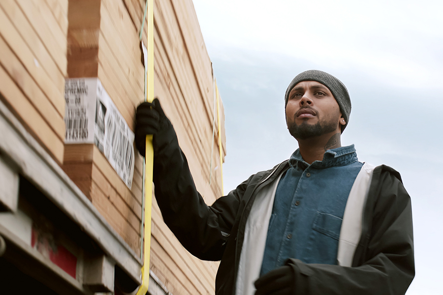 Truck driver securing lumber load with straps on a flatbed trailer.